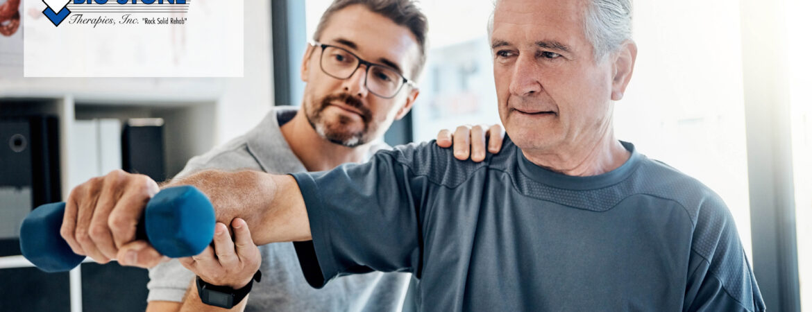 An elderly man in a blue shirt lifts a blue dumbbell, assisted by a younger man wearing glasses in a light room. The atmosphere is supportive and focused.