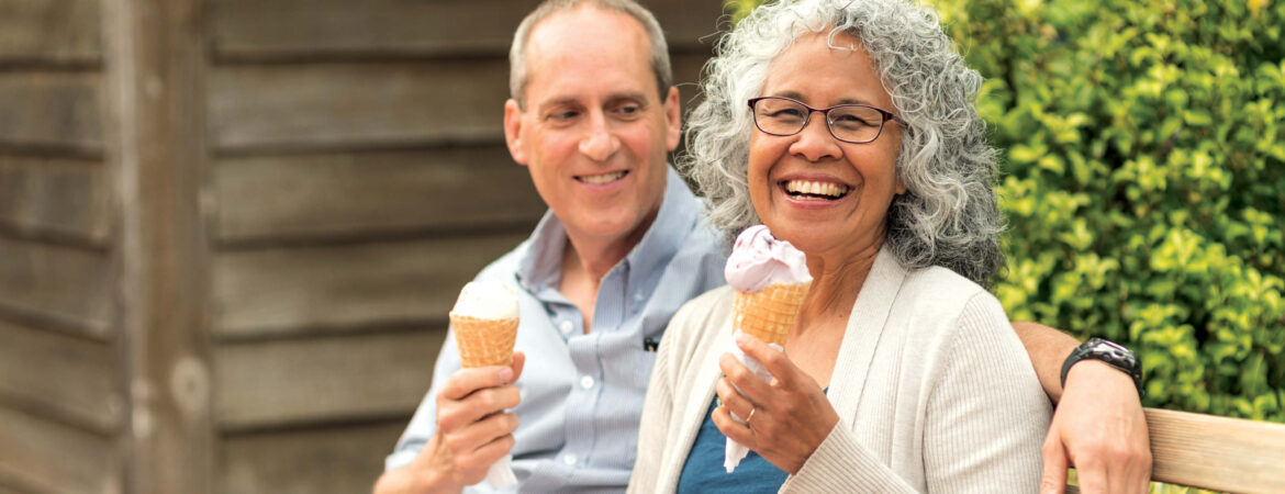 A smiling elderly couple sits on a bench, enjoying ice cream cones. The woman laughs joyfully, and the man looks at her warmly. Background features wooden siding and greenery.
