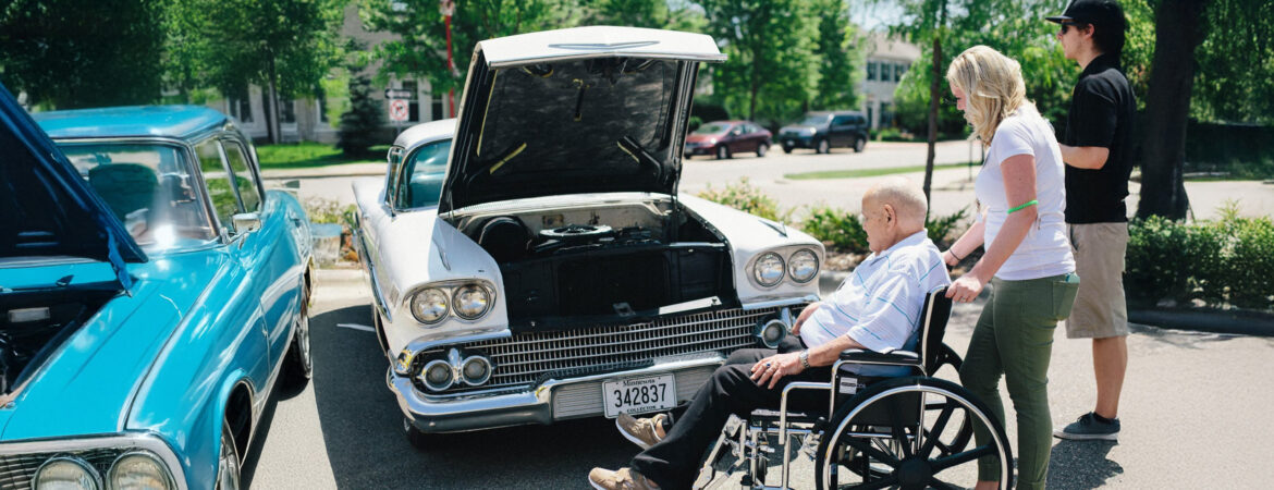 A man in a wheelchair views a classic car with its hood open at an outdoor car show. Two people stand nearby.