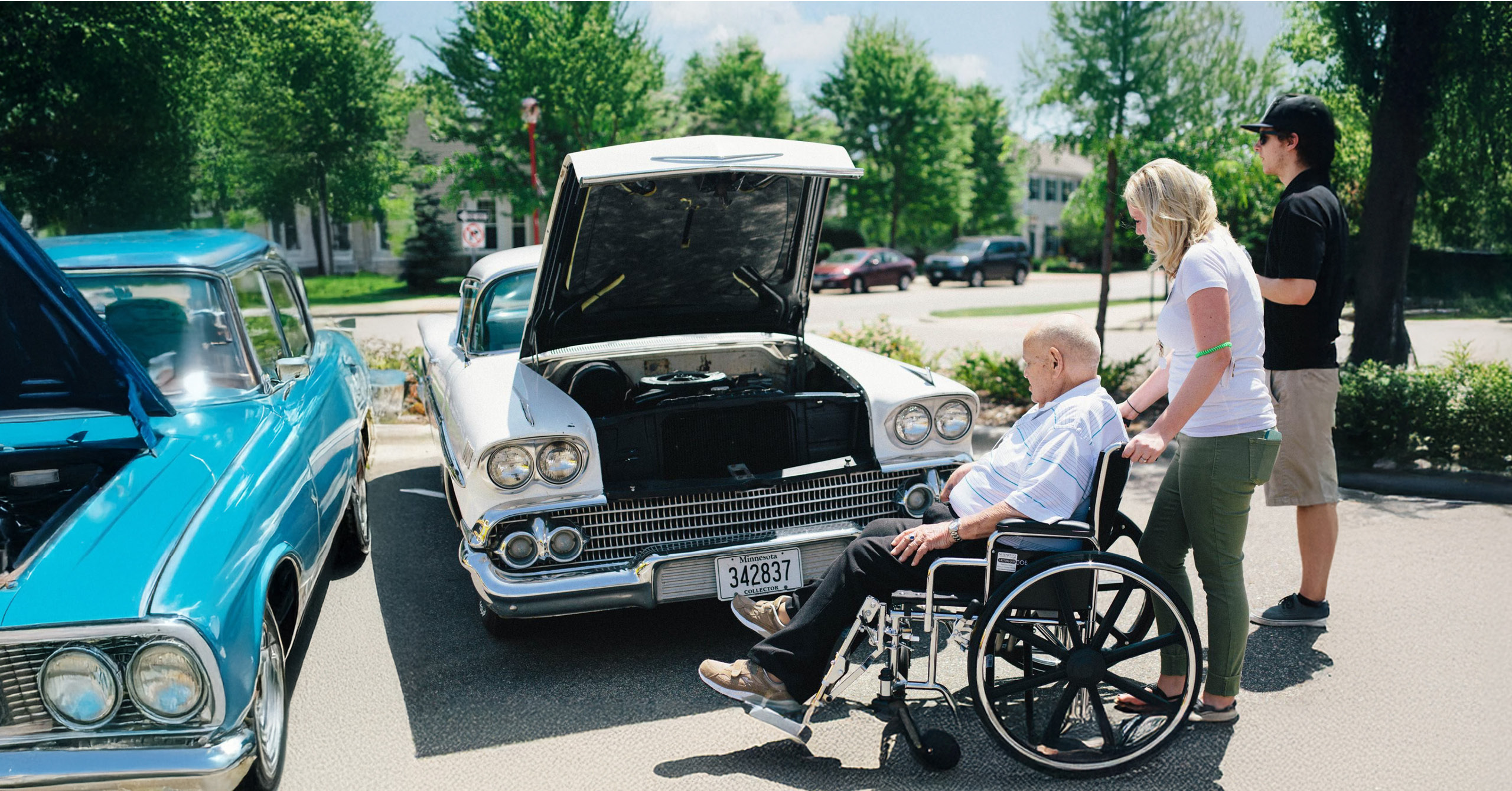 A man in a wheelchair views a classic car with its hood open at an outdoor car show. Two people stand nearby.